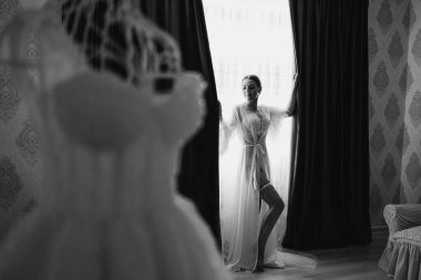 Beyaz elbiseli bir kadın perdenin önünde duruyor. Beyaz bir elbise ve beyaz bir duvak giyiyor.