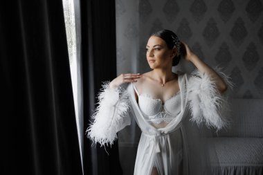 Üzerinde tüy olan beyaz elbiseli bir kadın pencerenin önünde duruyor. Kolye takıyor ve saçı topuz şeklinde. Zarafet ve incelik kavramı
