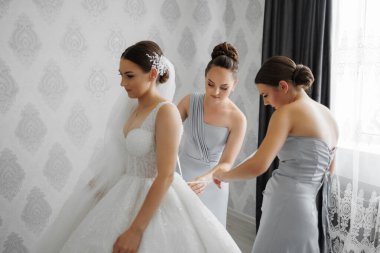 Üç kadın düğün için hazırlanıyor. Kadınlardan biri beyaz bir elbise giyiyor ve diğer ikisi mavi elbise giyiyor. Duvarın önünde duruyorlar, üzerinde bir desen var.