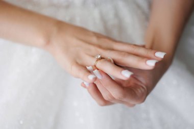 Bir kadın elinde bir yüzük tutuyor. Yüzük altın ve üzerinde elmas var. Kadın beyaz bir elbise giyiyor.