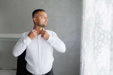 Beyaz gömlekli bir adam giyiniyor ve pencereden dışarı bakıyor. Rutin ve günlük hayat kavramı, adam sabah rutinine devam ederken işe gitmek için giyiniyor.