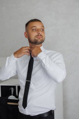 Bir adam kravatını bağlamaya hazırlanıyor. Beyaz gömlek ve siyah kravat takıyor.