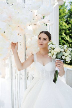 Bir kadın beyaz bir elbise giyiyor ve elinde bir buket beyaz çiçek tutuyor. Beyaz bir kemer yolunun önünde duruyor.