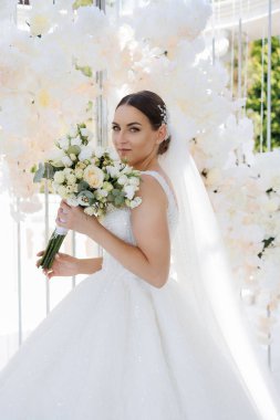 Bir gelin, elinde bir buket çiçek olan bir resim için poz veriyor. Beyaz bir elbise ve duvak takıyor. Çiçekler beyaz ve sarıdır ve bir vazoda düzenlenmişlerdir. Gelin gülümsüyor.