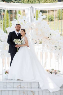 Bir gelin ve damat beyaz bir kemerin önünde poz veriyorlar. Gelin beyaz bir elbise giyiyor ve damat bir takım elbise giyiyor. Kemer beyaz çiçeklerle süslenmiş.