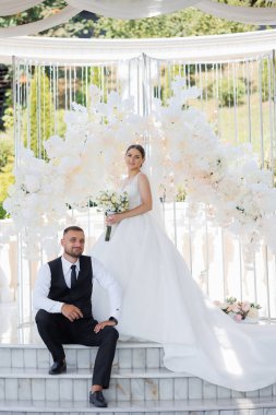 Gelin ve damat beyaz bir kemerin önünde fotoğraf çektirmek için poz veriyor. Gelin beyaz bir elbise giyiyor ve damat siyah bir yelek giyiyor. Kemer beyaz çiçeklerle süslenmiş.