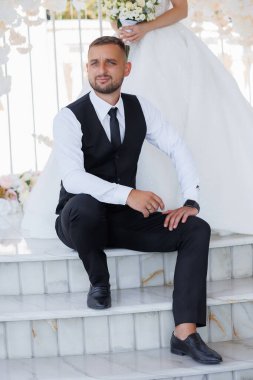 Siyah takım elbiseli ve beyaz gömlekli bir adam merdivenlerde oturuyor. Siyah bir kravat ve siyah bir yelek giyiyor. Adam gülümsüyor ve mutlu. Kutlama ve sevinç kavramı