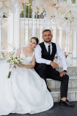 Bir gelin ve damat beyaz bir kemerin önünde poz veriyorlar. Gelin bir buket tutuyor ve damat kravat takıyor. Sahne mutlu ve kutlama dolu.