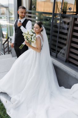 Bir gelin ve damat binanın dışında duruyor. Gelin bir buket çiçek tutuyor ve damat kravat takıyor.