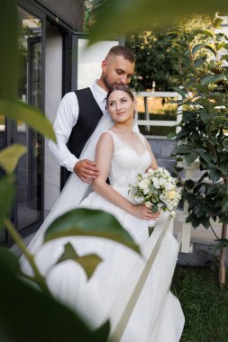 Bir gelin ve damat binanın önünde fotoğraf için poz veriyorlar. Gelin bir buket tutuyor ve damat elini tutuyor. Sahne mutlu ve romantik.