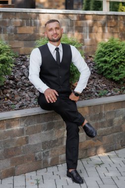 Takım elbiseli ve kravatlı bir adam tuğla bir duvara oturur. Siyah bir yelek ve beyaz bir gömlek giyiyor.