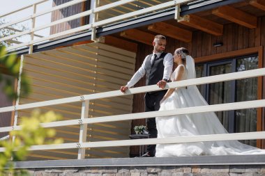 Gelin ve damat bir balkonda dikilip bahçeye bakıyorlar. Gelin beyaz bir elbise giyiyor ve damat siyah bir takım elbise giyiyor. Sahne romantik ve mutlu.