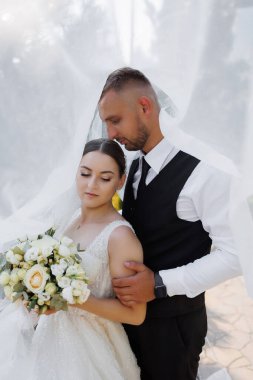 Bir gelin ve damat beyaz bir peçenin altında fotoğraf için poz veriyorlar. Gelin bir buket çiçek tutuyor ve damat siyah bir yelek giyiyor. Sahne romantik ve mutlu.