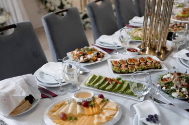 Bir tabak peynir, kraker, salata ve sandviç dahil çeşitli yiyecek ve içeceklerle dolu bir masa. Masa çoklu şarap kadehleri, çatallar, bıçaklar ve kaşıklarla dolu.