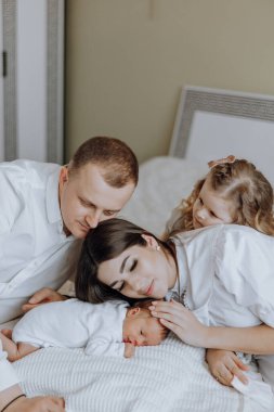 Dört kişilik bir aile, bir çift ve iki çocukları da dahil, yeni doğmuş bir bebeği yatakta kucaklıyor. Sahne sıcak ve sevgi dolu, çünkü aile bebeğe şefkat ve ilgi gösteriyor.