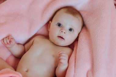 Bir bebek gözleri açık pembe bir battaniyenin üzerinde yatıyor. Bebek kameraya bakıyor ve merak ediyor.