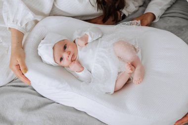 Beyaz bir battaniyeyle beyaz bir yatakta yatan bir bebek var. Bebek beyaz bir elbise ve beyaz bir şapka giyiyor. Bebek kameraya bakıyor.