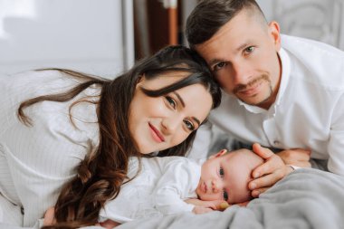 Genç bir çift yataklarında bir bebek tutuyor. Bebek beyaz bir kıyafet giyiyor. Sahne sıcak ve sevgi dolu, çünkü çift yeni çocuklarını kucaklıyor.