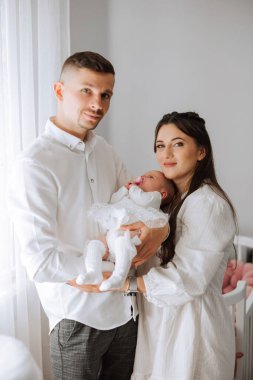 Beyaz bir odada bir erkek ve bir kadın bir bebek tutuyor. Bebeğin beyaz bir elbise giyiyor ve pembe bir burnu var. Sahne mutlu ve neşeli. Aile yeni çocuklarının doğumunu kutluyor.