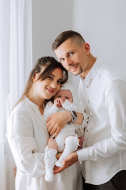 Genç bir çift kucağında bir bebek tutuyor. Bebek beyaz bir elbise ve beyaz bir şapka giyiyor. Aile bir resim için poz veriyor ve görüntünün havası mutlu ve sevgi dolu.