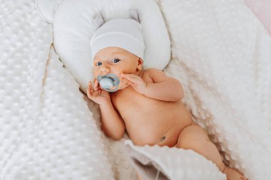 Bir bebek ağzında bir emzik ile bir yatakta yatıyor. Bebek beyaz bir şapka takıyor ve kameraya bakıyor.