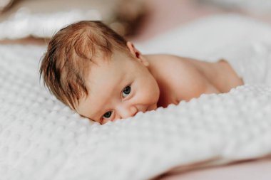 Bir bebek başı yana eğilmiş bir şekilde bir yatağın üzerinde yatıyor. Bebeğin yüzünde bir gülümseme var ve kameraya bakıyor. Bebeğin memnuniyeti gibi görünen sıcaklık ve mutluluk kavramı.