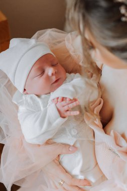 Bir bebek pembe elbiseli bir kadın tarafından tutuluyor. Bebek beyaz bir şapka ve beyaz bir kıyafet giyiyor. Kadın bebeği göğsüne yakın tutuyor ve bebek uyuyor.