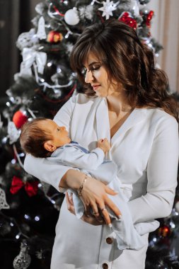 Bir kadın kucağında bir bebek tutuyor. Bebek mavi bir gömlek giyiyor. Kadın beyaz bir elbise giyiyor. Sahne bir Noel ağacının önünde geçiyor.
