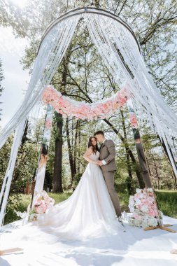 Bir gelin ve damat ormanda beyaz bir gölgeliğin altında duruyor. Gelin beyaz bir elbise giyiyor ve damat bir takım elbise giyiyor. Sahne romantik ve samimi.