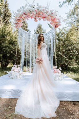 Beyaz bir kemerin önünde elinde pembe çiçeklerle bir gelin duruyor. O, elinde bir buket çiçek tutuyor. Sahne bir bahçede geçiyor, arka planda ağaçlar ve çalılar var.