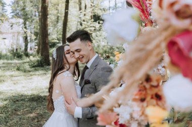 Bir gelin ve damat ormanda sarılıyor. Gelin beyaz bir elbise giyiyor ve damat da takım elbise giyiyor.