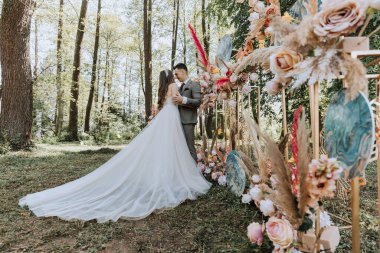 Bir gelin ve damat çiçek kemerinin önünde öpüşüyorlar. Gelin beyaz bir elbise giyiyor ve damat bir takım elbise giyiyor. Kemer çiçeklerle süslenmiş ve mavi-beyaz bir tasarımı var.