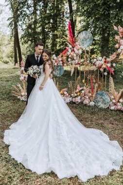 Bir gelin ve damat çiçek kemerinin önünde poz veriyorlar. Gelin beyaz bir elbise giyiyor ve damat bir takım elbise giyiyor. Kemer çiçeklerle süslenmiş ve mavi bir kemeri var.