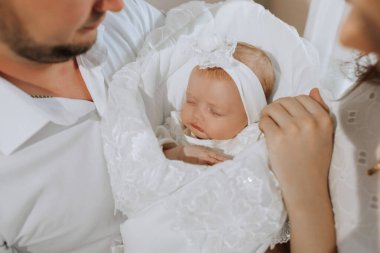 Beyaz bir battaniyenin üzerinde bir bebek uyuyor. Bebeği bir erkek ve bir kadın tutuyor.