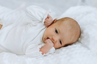 Bir bebek beyaz bir battaniyenin üzerinde yatıyor, başı yana dönük. Bebek kameraya bakıyor ve ağzı açık vaziyette. Masumiyet ve merak kavramı