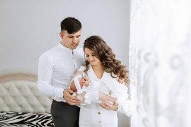 Beyaz bir odada bir çift bir bebek tutuyor. Bebek beyaz bir kıyafet giyiyor. Odada zebra desenli yatak örtüsü var.