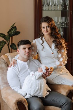 Kollarında bir bebekle kanepede oturan genç bir çift. Bebek beyaz bir elbise giyiyor ve çift beyaz giyinmiş. Sahne sıcak ve sevgi dolu.