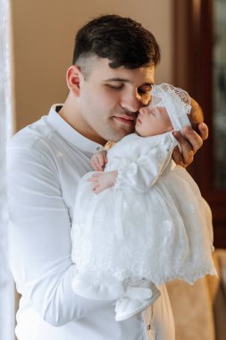Bir adam kucağında bir bebek tutuyor. Bebek beyaz bir elbise ve beyaz bir saç bandı takıyor. Adam bebeğe nazikçe bakıyor.