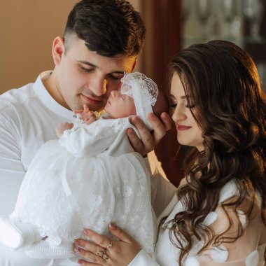 Genç bir çift beyaz elbiseli bir bebek tutuyor. Bebek beyaz bir saç bandı takıyor. Aile gülümsüyor ve o mutlu.