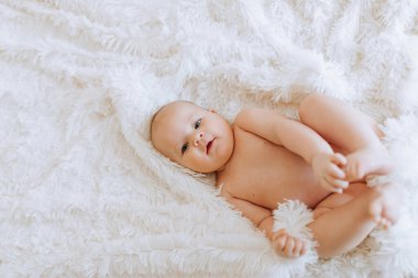Beyaz bir battaniyenin üzerinde bir bebek yatıyor. Bebek gülümsüyor ve kameraya bakıyor. Bebeğin ilgi odağı olduğu sahne huzurlu ve sıcak.