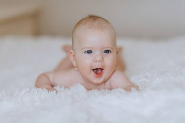 Yüzünde bir gülümsemeyle beyaz bir battaniyeye uzanmış bir bebek. Bebek gülümsüyor ve komik bir yüz ifadesi takınıyor. Bu da resme mutlu ve neşeli bir hava veriyor.