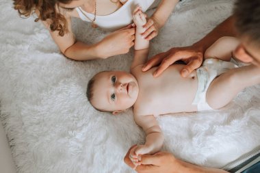 Bir bebek, iki yetişkinin kolunu tuttuğu bir yatakta yatıyor. Bebek gülümsüyor ve ilginin tadını çıkarıyor. Yetişkinler ve bebek arasındaki sıcaklık ve sevgi kavramı.