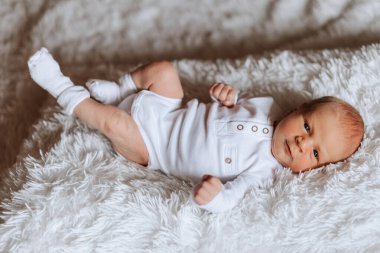 Beyaz bir battaniyenin üzerinde bir bebek yatıyor. Bebek beyaz çorap ve beyaz tulum giyiyor.