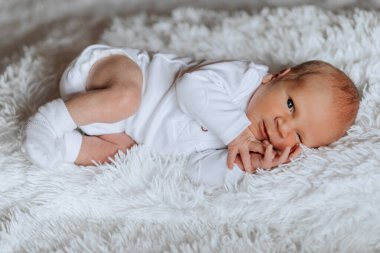 Beyaz bir battaniyenin üzerinde bir bebek yatıyor. Bebek beyaz bir tulum giyiyor ve beyaz bir çorabı var. Bebek gülümseyerek kameraya bakıyor.