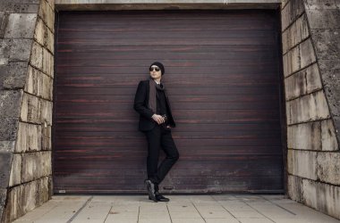 Takım elbiseli ve şapkalı bir adam garaj kapısının önünde duruyor. Güneş gözlüğü ve kravat takıyor.