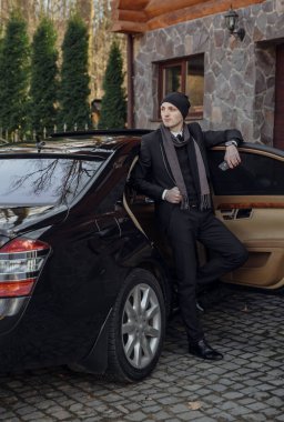 Takım elbiseli ve şapkalı bir adam siyah bir arabanın yanında duruyor. Elinde bir içki var ve birini bekliyor.