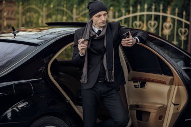 Siyah takım elbiseli ve şapkalı bir adam arabanın arkasında duruyor. Elinde bir cep telefonu var ve kontrol ediyor.