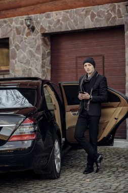 Siyah takım elbiseli ve şapkalı bir adam siyah bir arabanın önünde duruyor. Elinde bir cep telefonu var ve birini bekliyor.
