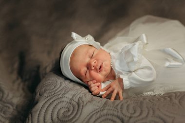 Yeni doğmuş bir bebek, beyaz bir elbise ve kafasında beyaz bir fiyonk ile bir yatakta huzur içinde uyuyor. Bebeğin yüzü göğsünde dinleniyor ve minicik eli alnında dinleniyor.