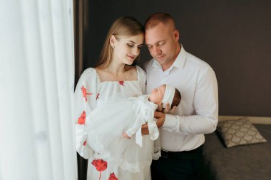 Genç bir çift beyaz elbiseli bir bebek tutuyor. Bebek beyaz bir saç bandı takıyor. Aile bir pencerenin önünde duruyor.